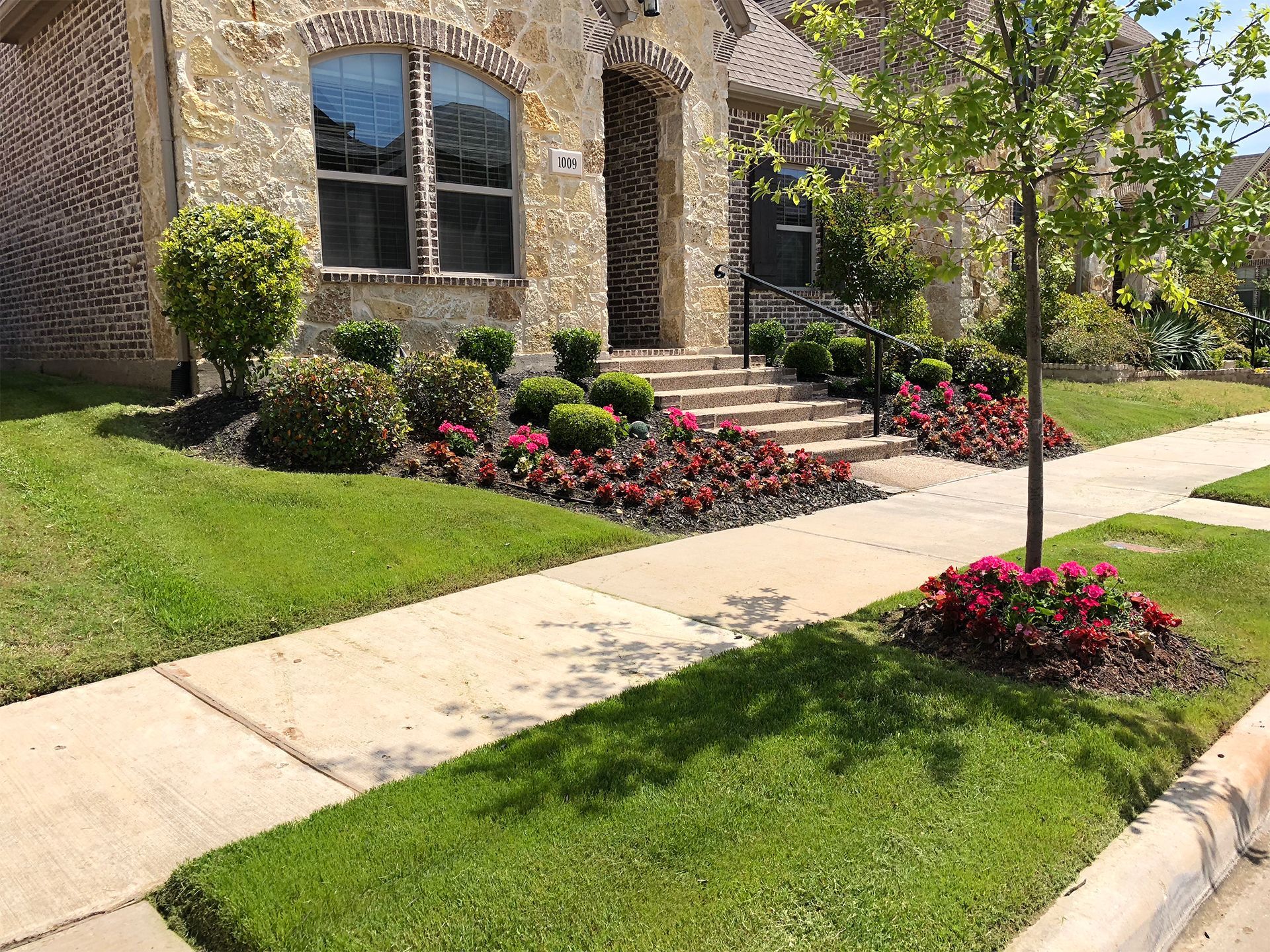 Suburban house facade with stone walls, front steps, walkway, and colorful flower beds