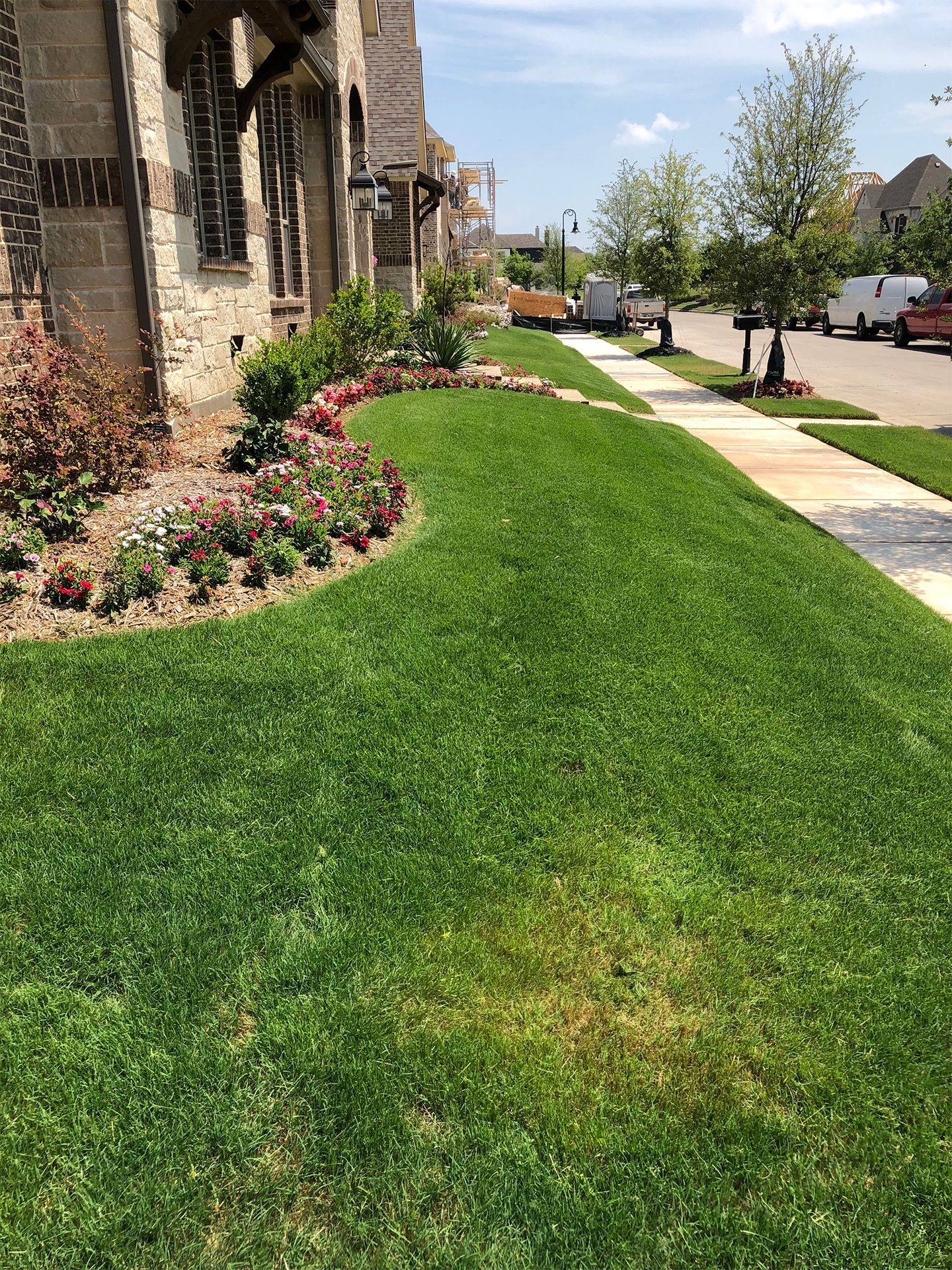 Sunlit sidewalk beside a stone building, with neatly edged green lawn and flower beds along a quiet street.