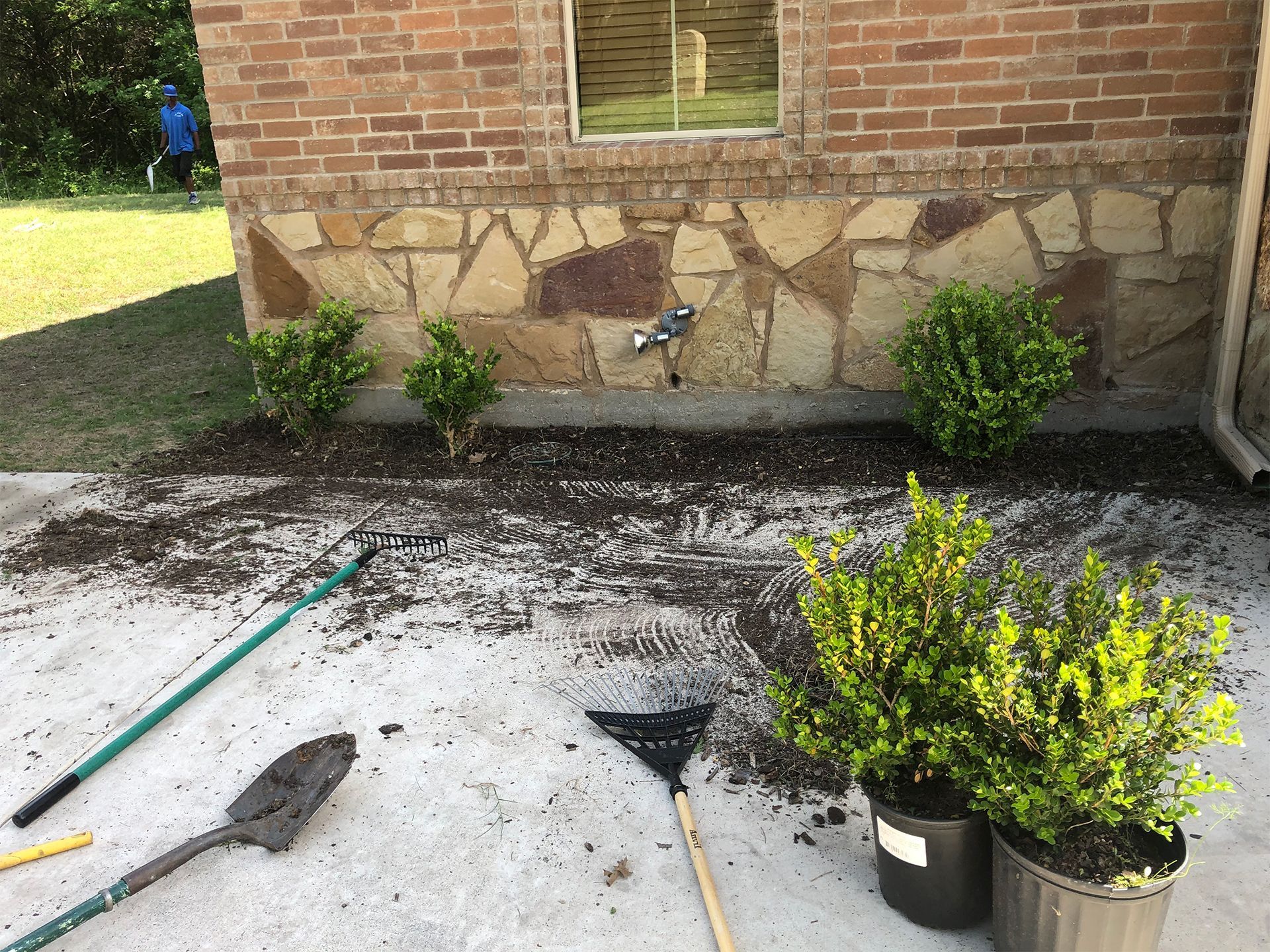 Backyard patio with garden tools, potted shrubs, and a brick house wall with decorative stonework.