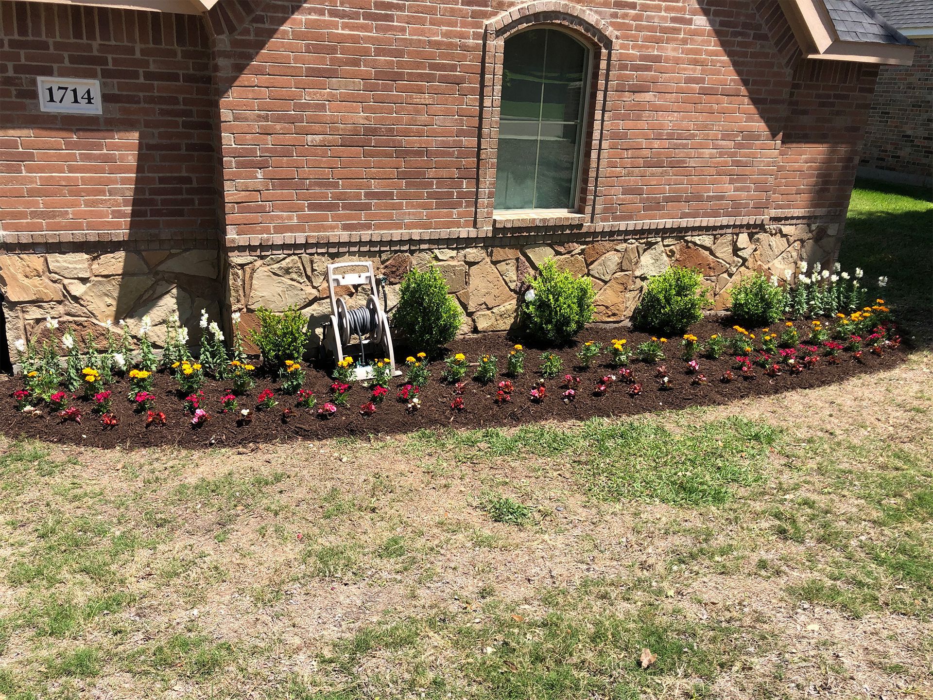 Brick house front with flower bed, small statues, and a sunlit lawn