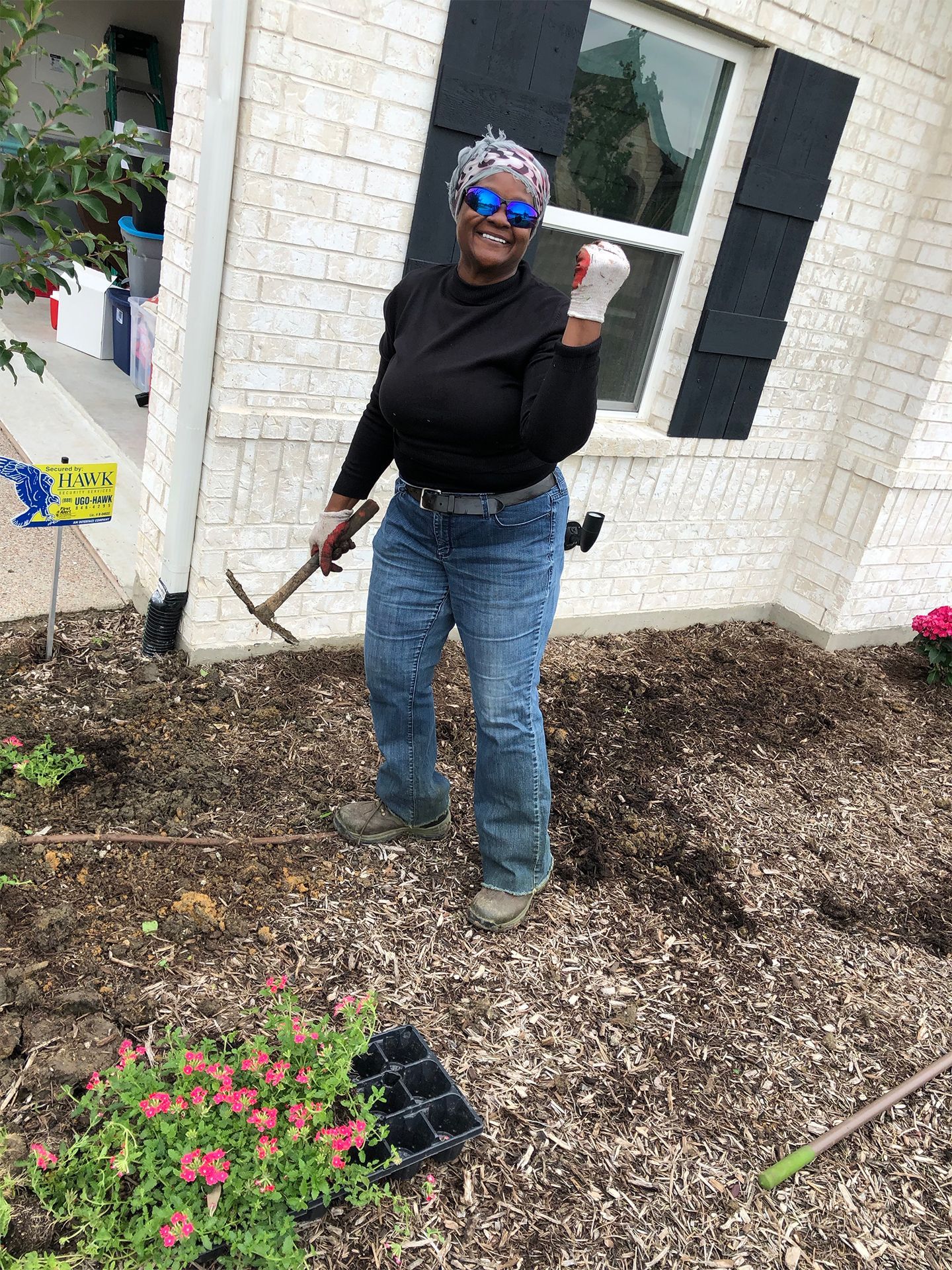 Woman standing in a garden holding a trowel, wearing sunglasses, near a white brick house.