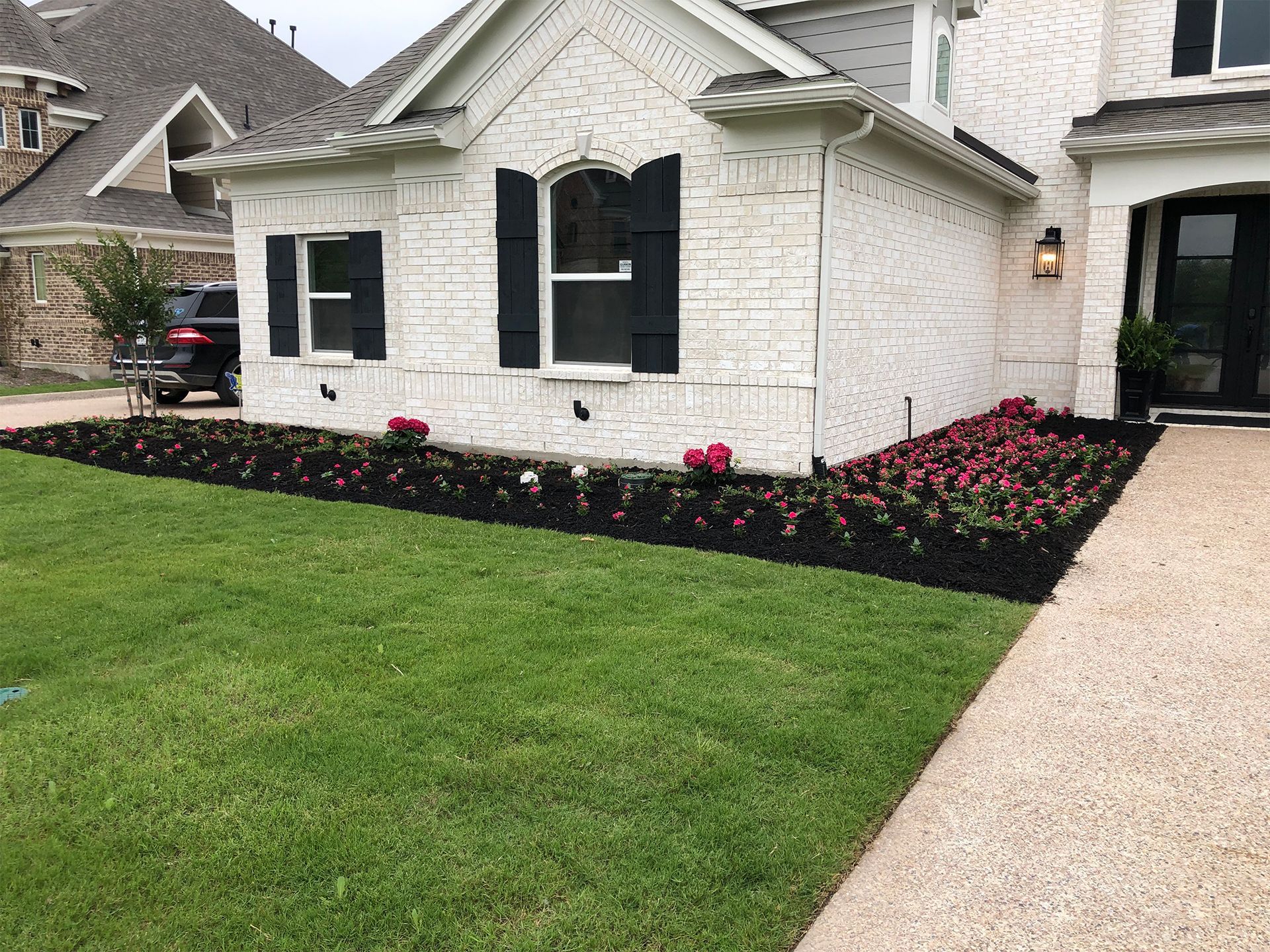 White house with black shutters, green lawn, pink flowers, and a gravel walkway in front