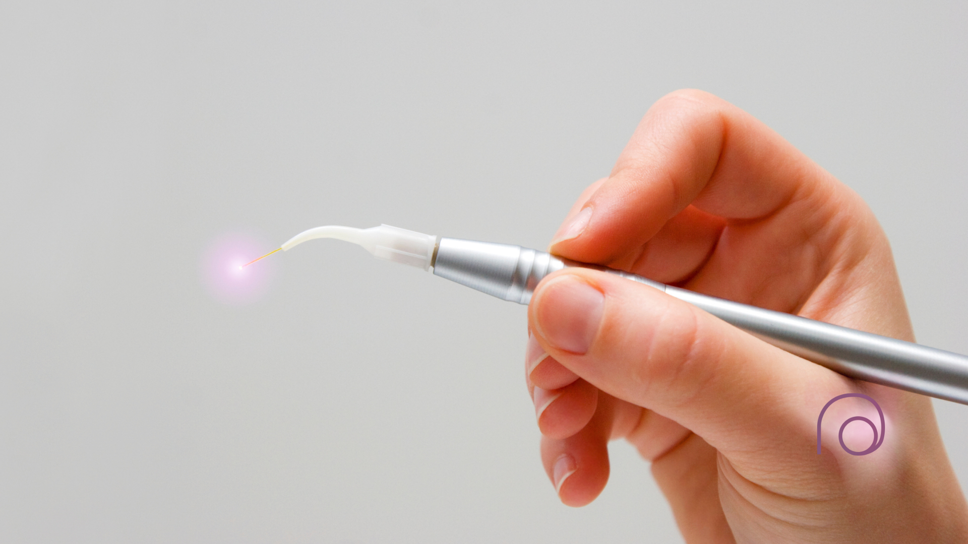 A hand holds a silver dental curing light with a glowing, light-emitting tip against a plain gray background.