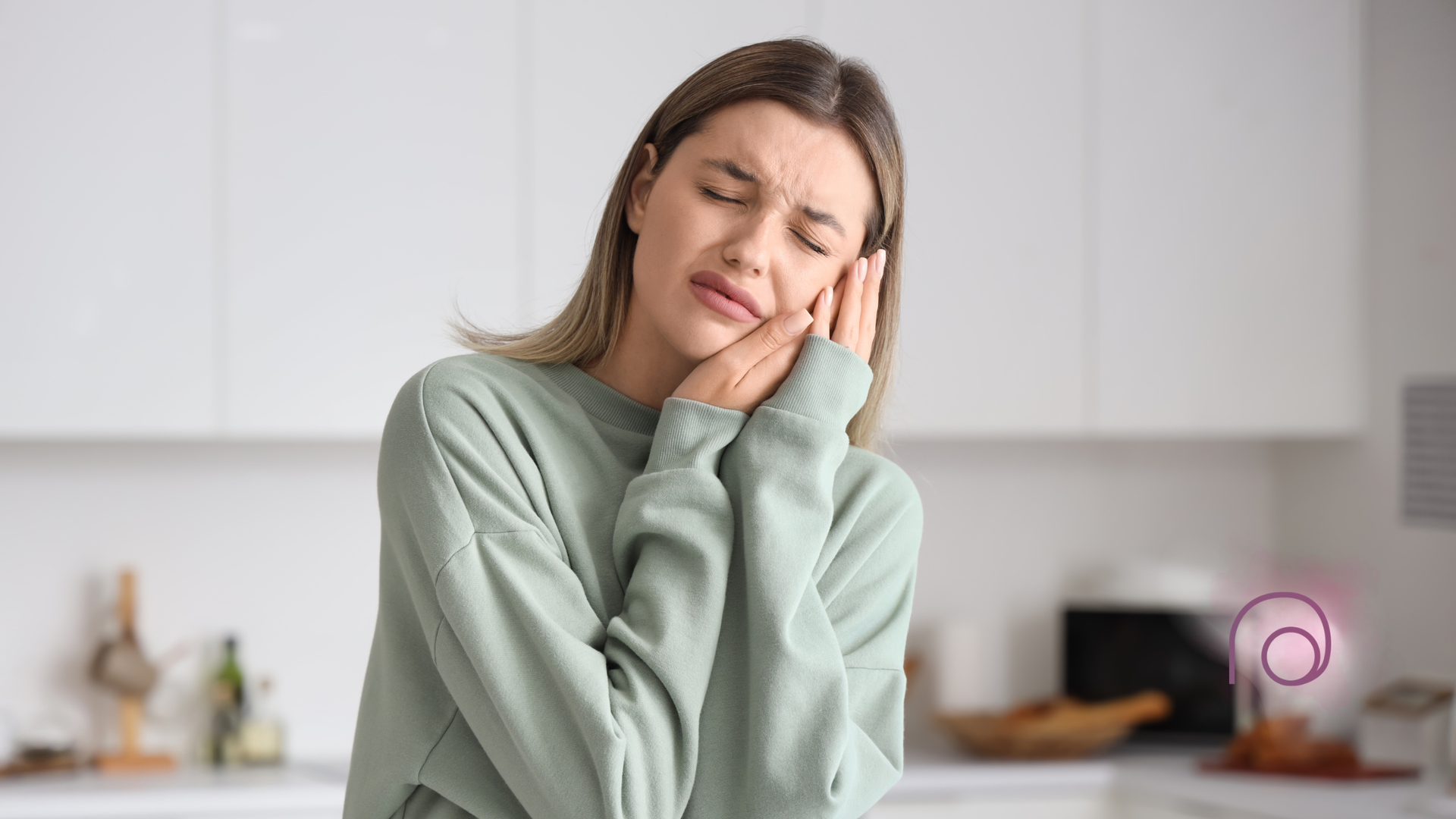 Woman in kitchen clutching cheek, eyes closed, appearing to have a toothache.