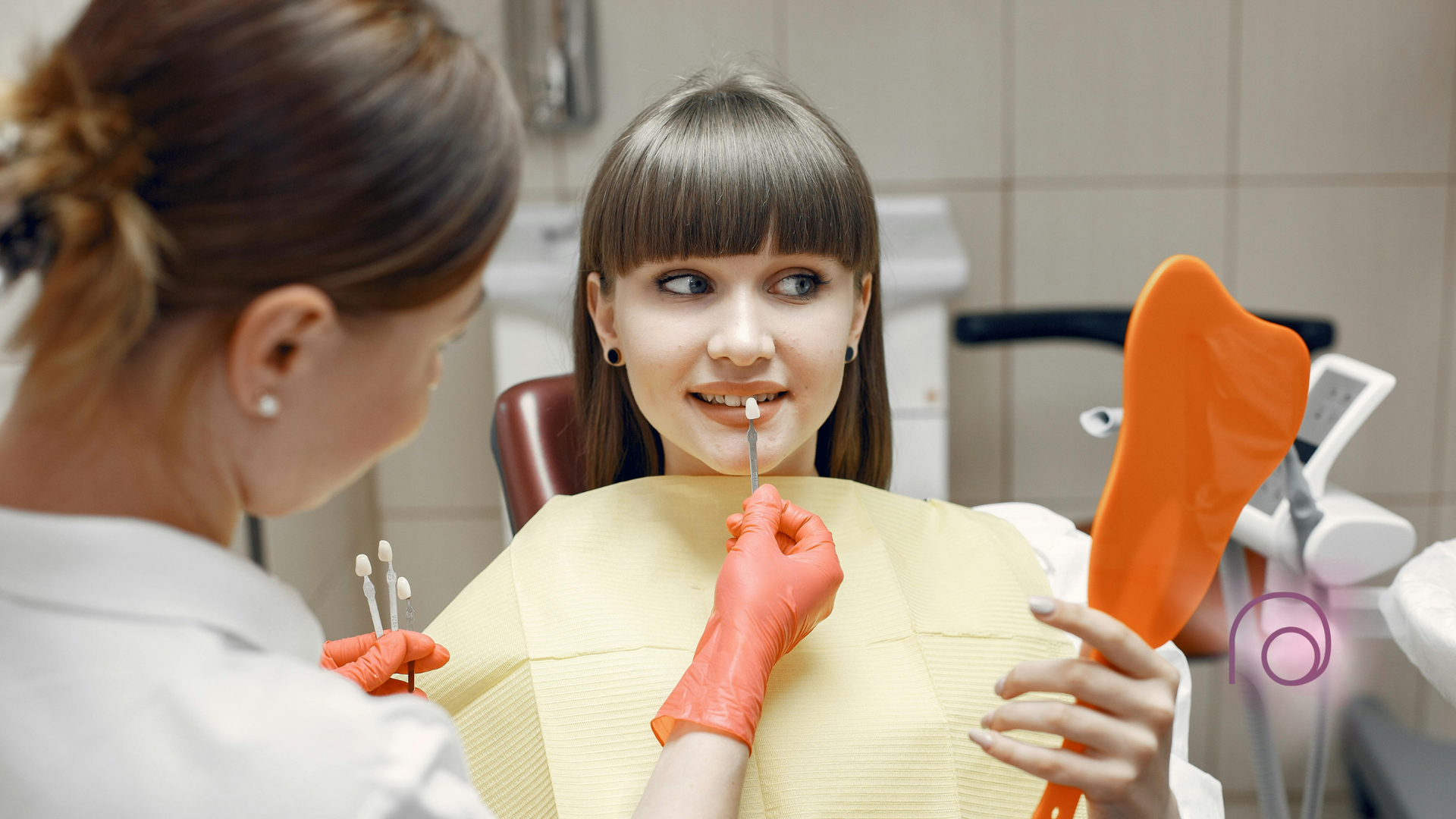 Woman at dentist comparing teeth shade with orange mirror, doctor holding shade guide.