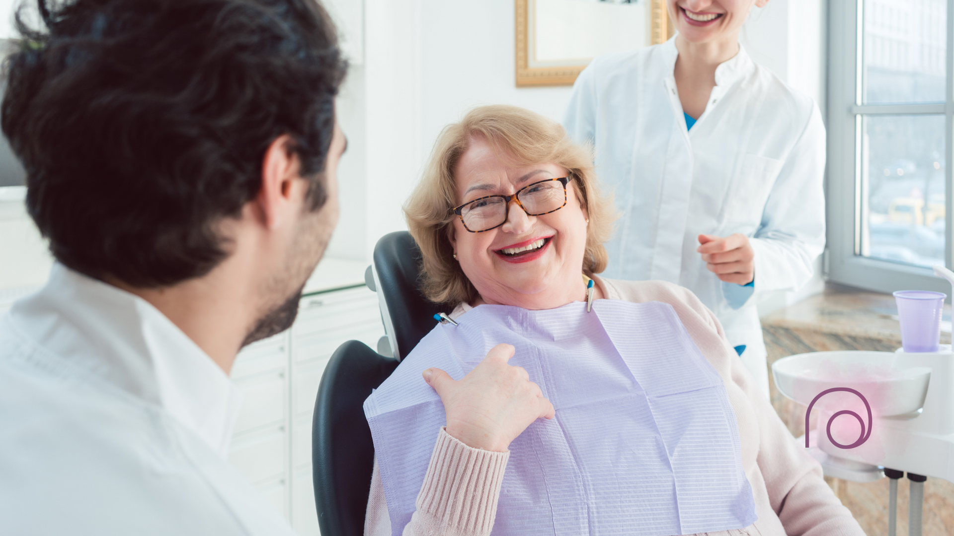 A smiling patient in a dental chair talks to a dentist while another staff member stands nearby in a bright office.