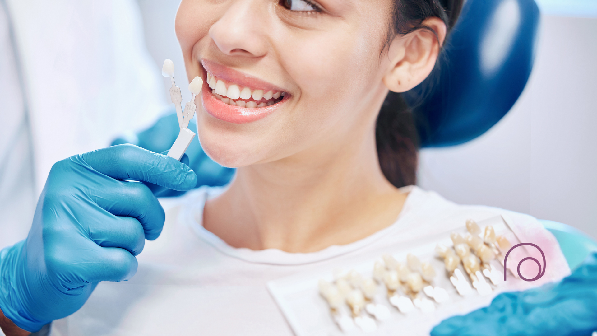A dentist in blue gloves holds a tooth shade guide near a patient's mouth to compare color for dental treatment.
