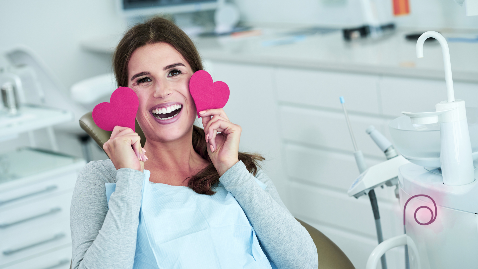 A woman is sitting in a dental chair holding two pink hearts in front of her eyes.