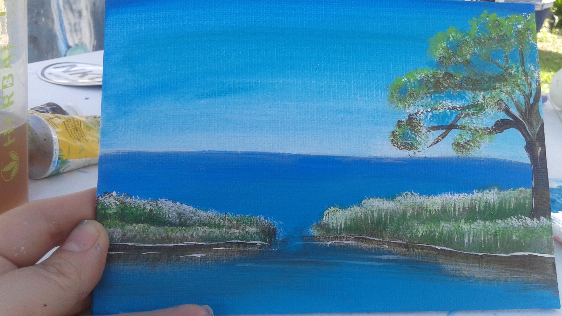 A person is holding a small painting of a lake and a tree.