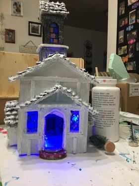 A cardboard gingerbread house model is sitting on a table next to a bottle of glue.