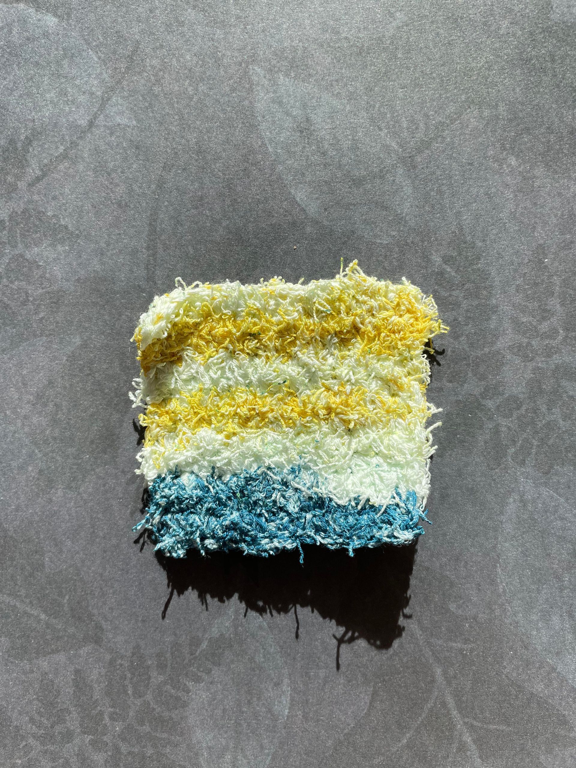 A handmade facial scrubbing square made from  yellow and blue yarn is sitting on a table.