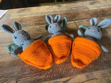 Three crocheted rabbits are sitting inside of carrots on a wooden table.