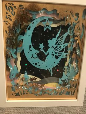 A paper craft scene with a fairy sitting on a crescent moon with a star in her hand.