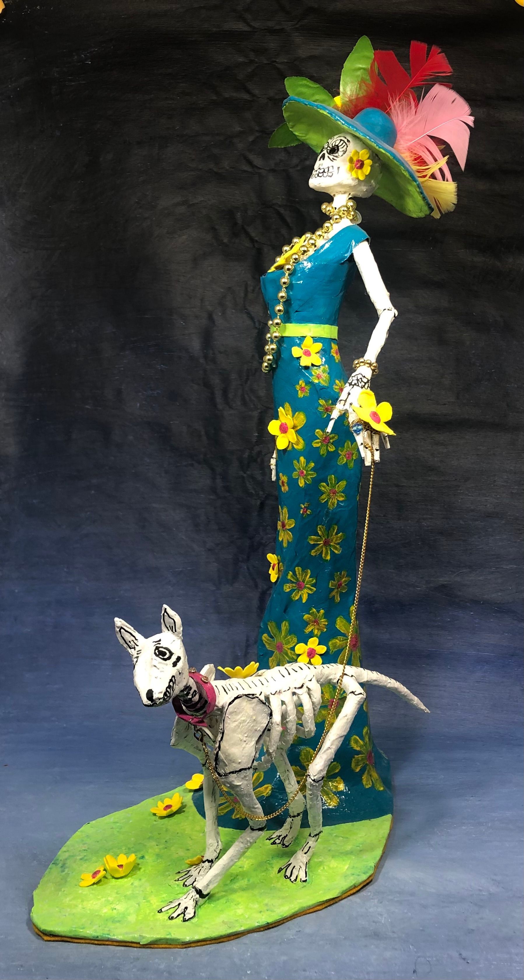A handmade skeleton figurine posing in a a tight fitting long teal gown with yellow flowers and a skeleton dog at her feet