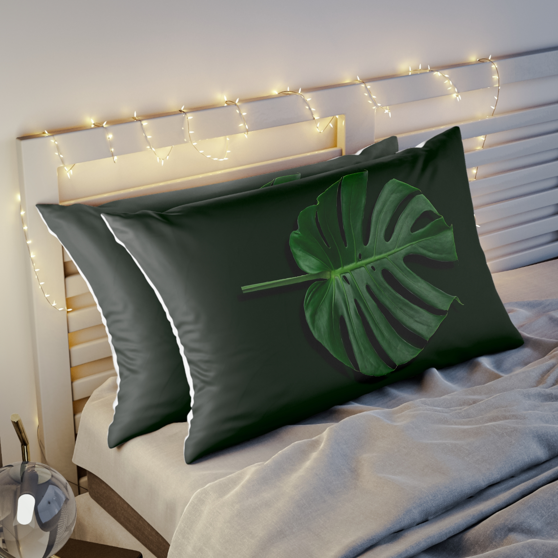 Giant Green Monstera Leaf on black background made into a pillow sham sitting on a bed with beige sheets andfairy lights