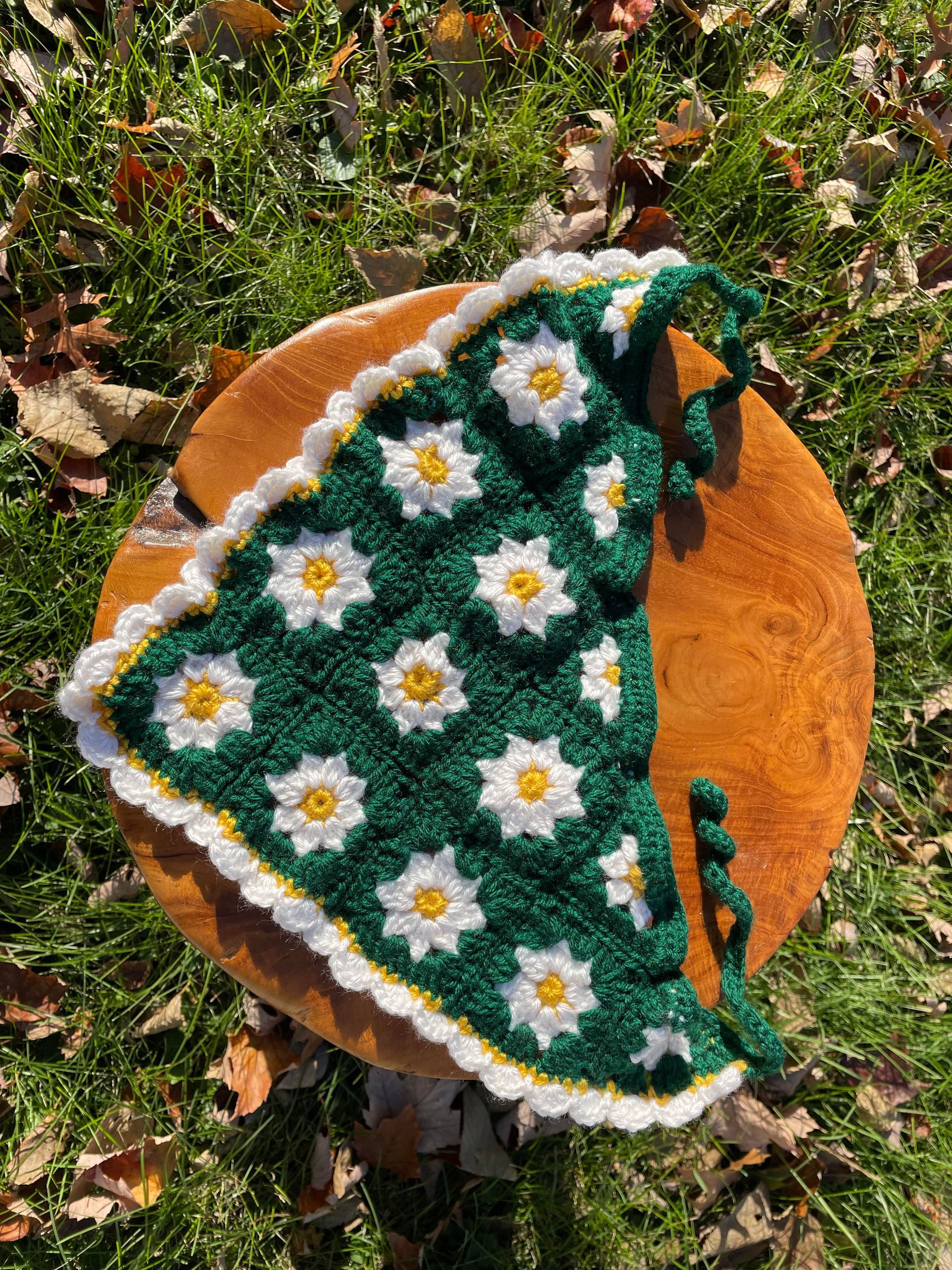 A green and white crocheted scarf is sitting on a wooden stool in the grass.