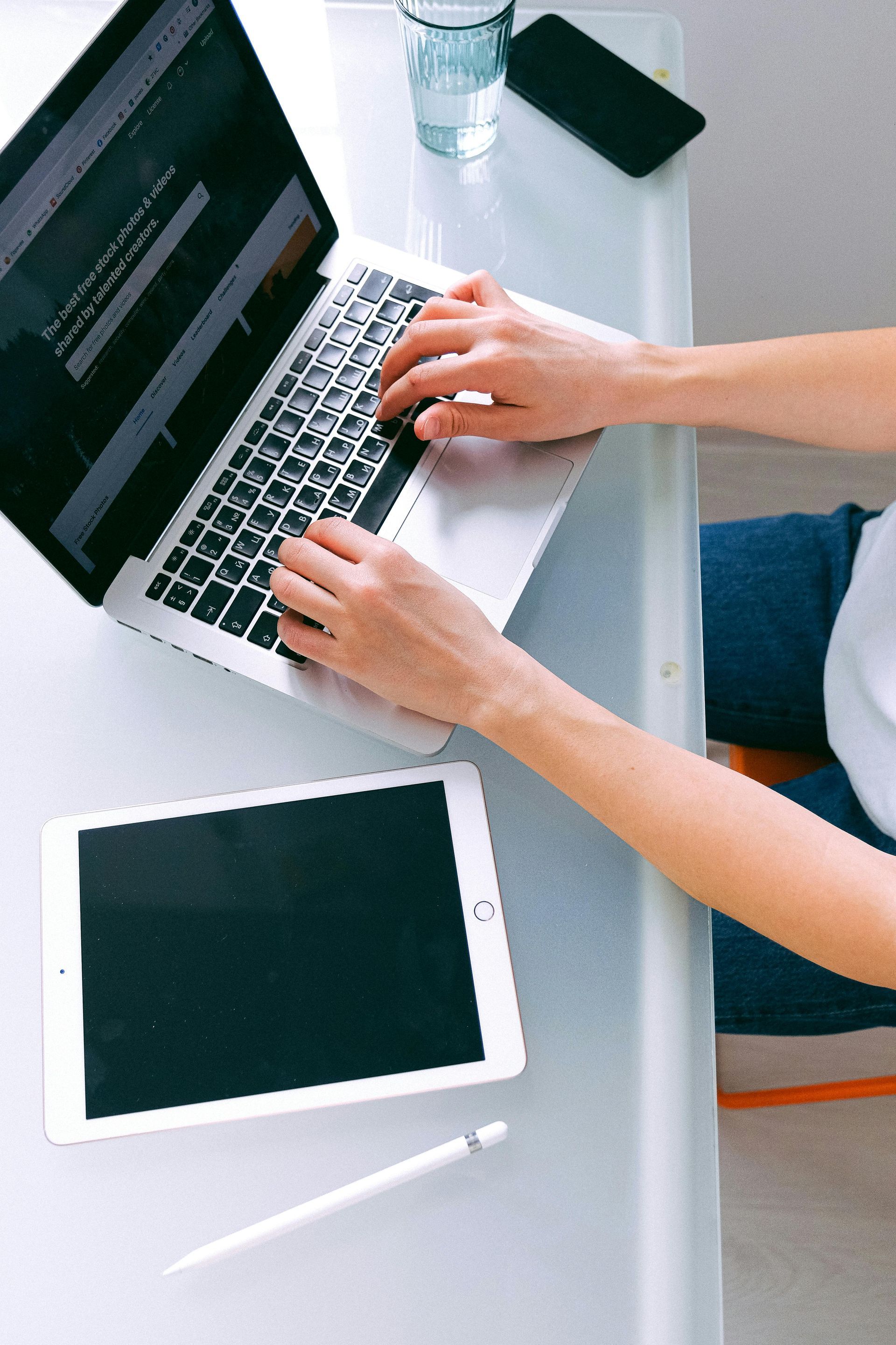 Person typing on a laptop at a desk with a tablet, phone, water glass, and pen.