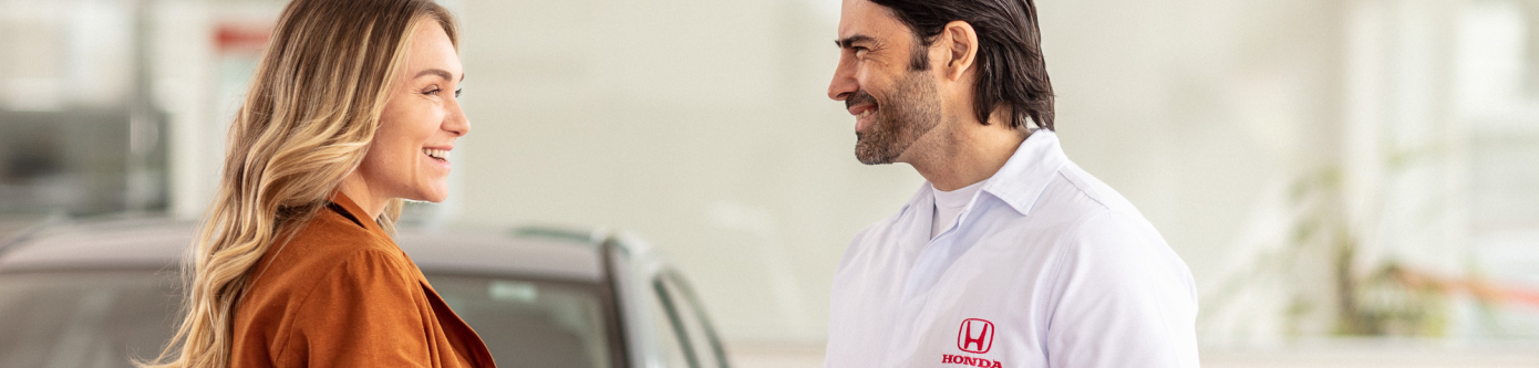 Uma mulher e um homem com uniforme claro interagem sorrindo um para o outro em uma concessionária de carros bem iluminada.