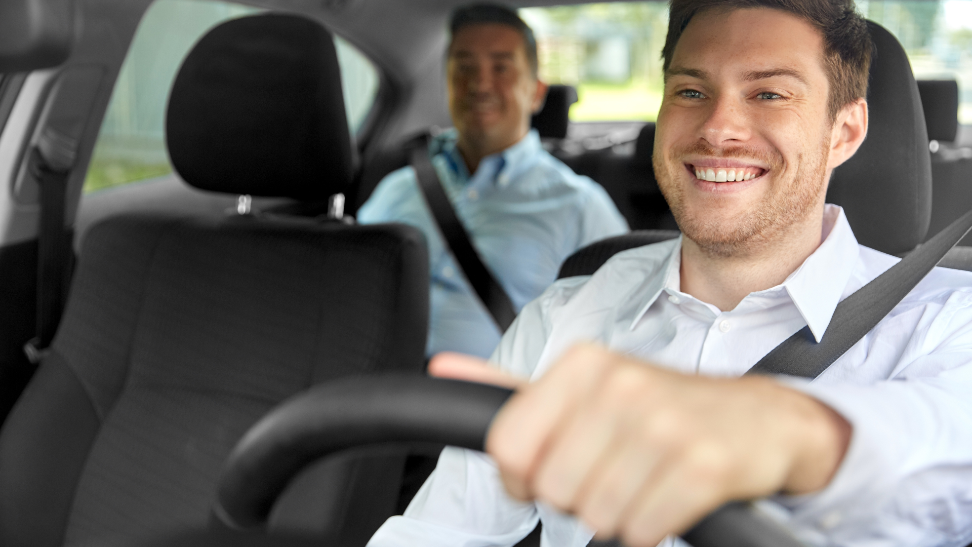 Um motorista sorridente, vestindo uma camisa clara, dirige um carro enquanto um passageiro está sentado no banco de trás.