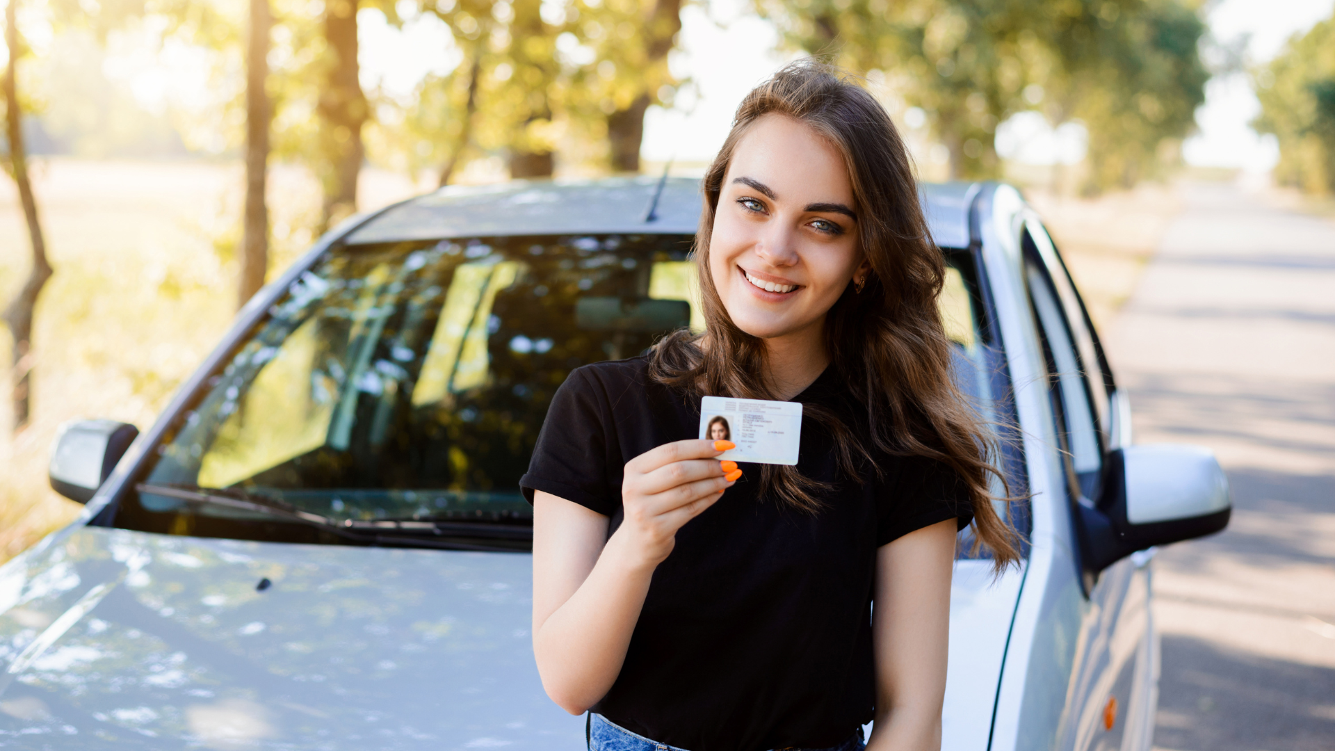 Uma pessoa sorrindo e segurando uma carteira de motorista em frente a um carro prata estacionado em uma rua ensolarada.