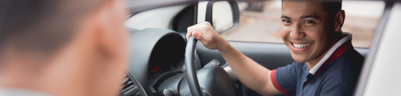 Um motorista sorridente, vestindo uma camisa polo azul, olha para um passageiro enquanto está sentado no banco do motorista de um carro.