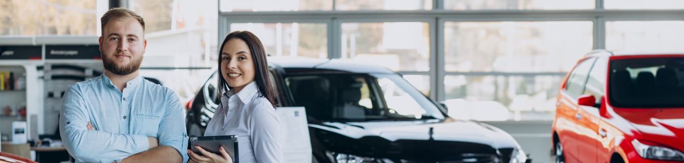 Um homem e uma mulher sorriem em uma concessionária de carros bem iluminada, com veículos pretos e vermelhos visíveis ao fundo.