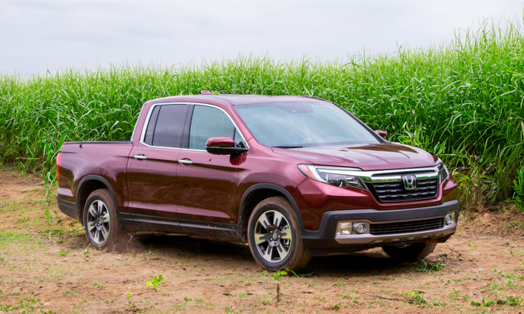 Uma caminhonete Honda Ridgeline bordô estacionada em uma estrada de terra ao lado de um campo de grama alta e verde.
