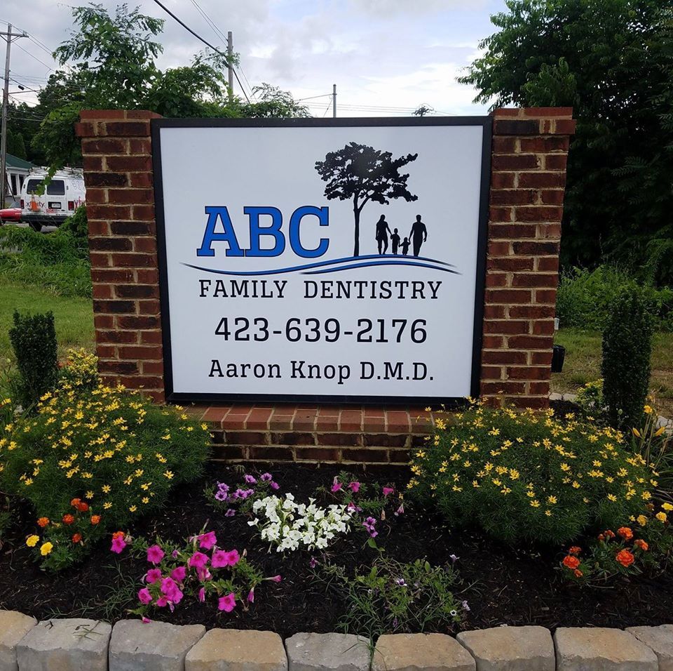 Sign for ABC Family Dentistry with logo, contact info, and name; surrounded by flowers and brick.