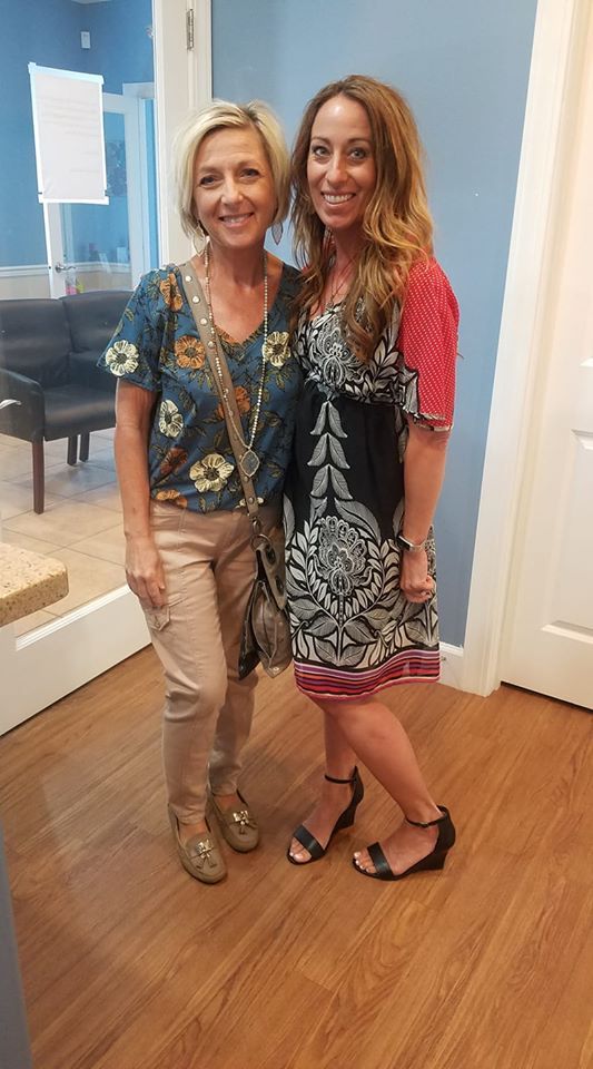 Two women smiling, posing indoors. Blonde with patterned top, tan pants, other in patterned dress and sandals.