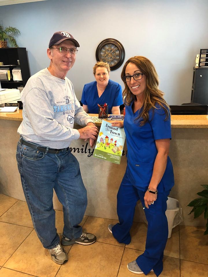 Man and two women in scrubs at a reception desk, holding a book. Blue scrubs, casual clothes, interior.