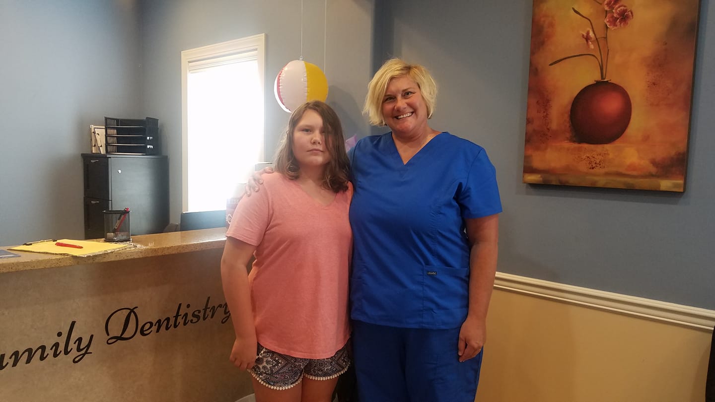 Woman in blue scrubs and girl pose at a dental office reception desk.