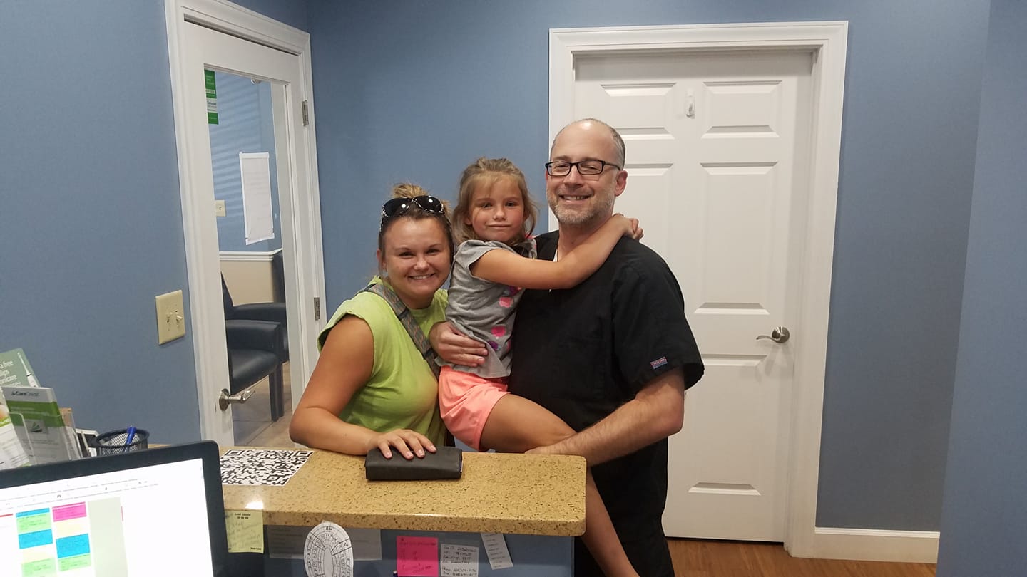 Family in dentist's office: woman, child, and man (dentist) smiling at the reception desk.
