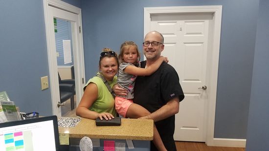 Family in dentist's office: woman, child, and man (dentist) smiling at the reception desk.