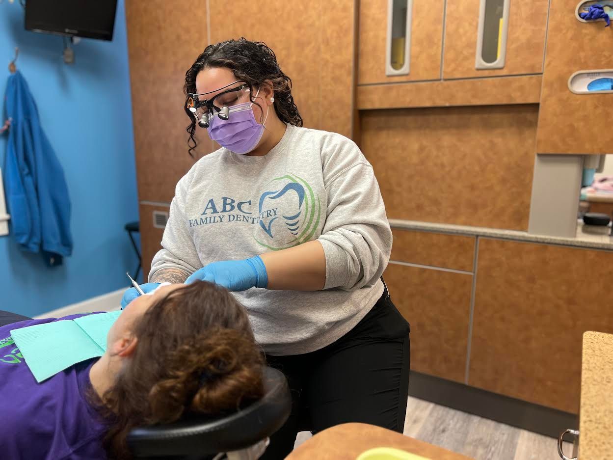 Dentist in mask and gloves examining a patient's teeth in an exam room.