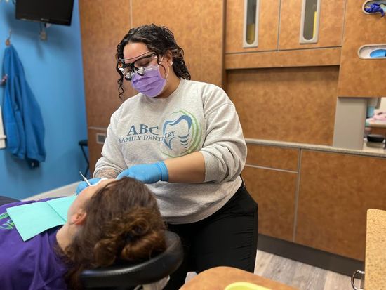 Dentist in mask and gloves examining a patient's teeth in an exam room.
