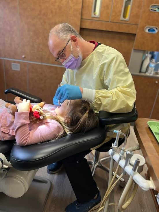 Dentist examining a young child's teeth in a dental chair. Both wear masks; the dentist wears gloves and a gown.