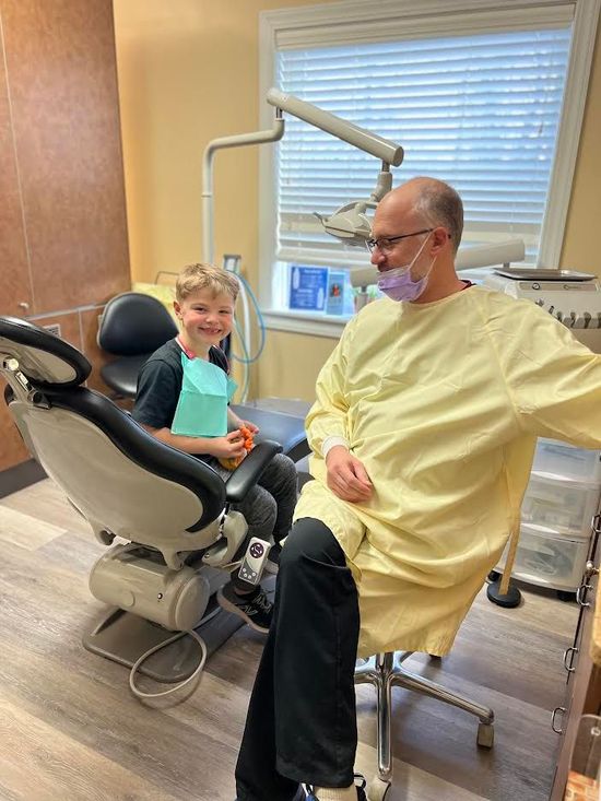 A young boy smiles in a dental chair next to a dentist wearing a mask and gown in a dental office.
