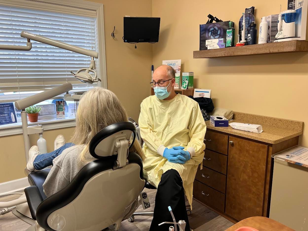 Dentist in mask and gown, sits with patient in dental chair, in a dental office.