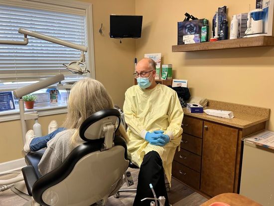 Dentist in mask and gown, sits with patient in dental chair, in a dental office.