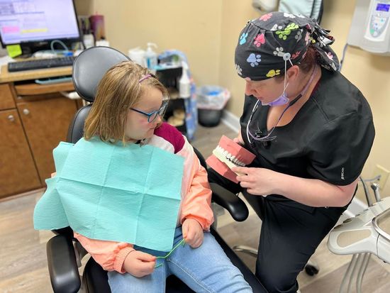 A dentist shows a young girl a tooth model. Both are in a dental office.