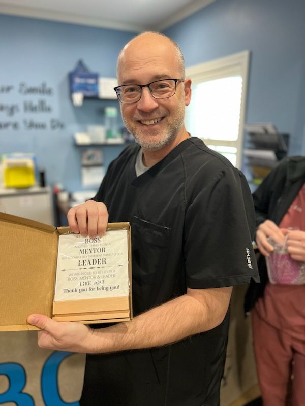 Man in black scrubs holding a framed award, smiling. Office setting, blue and brown tones.