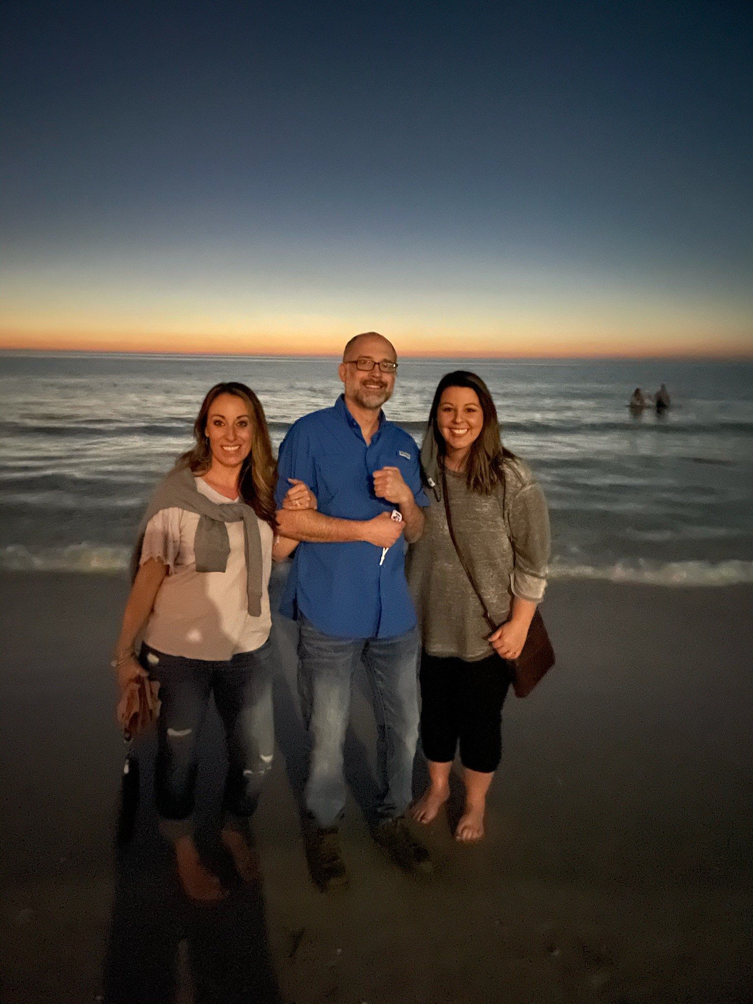 Three people standing on a beach at sunset: two women and a man, all smiling.