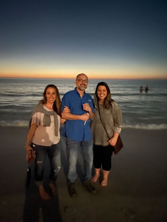Three people standing on a beach at sunset: two women and a man, all smiling.