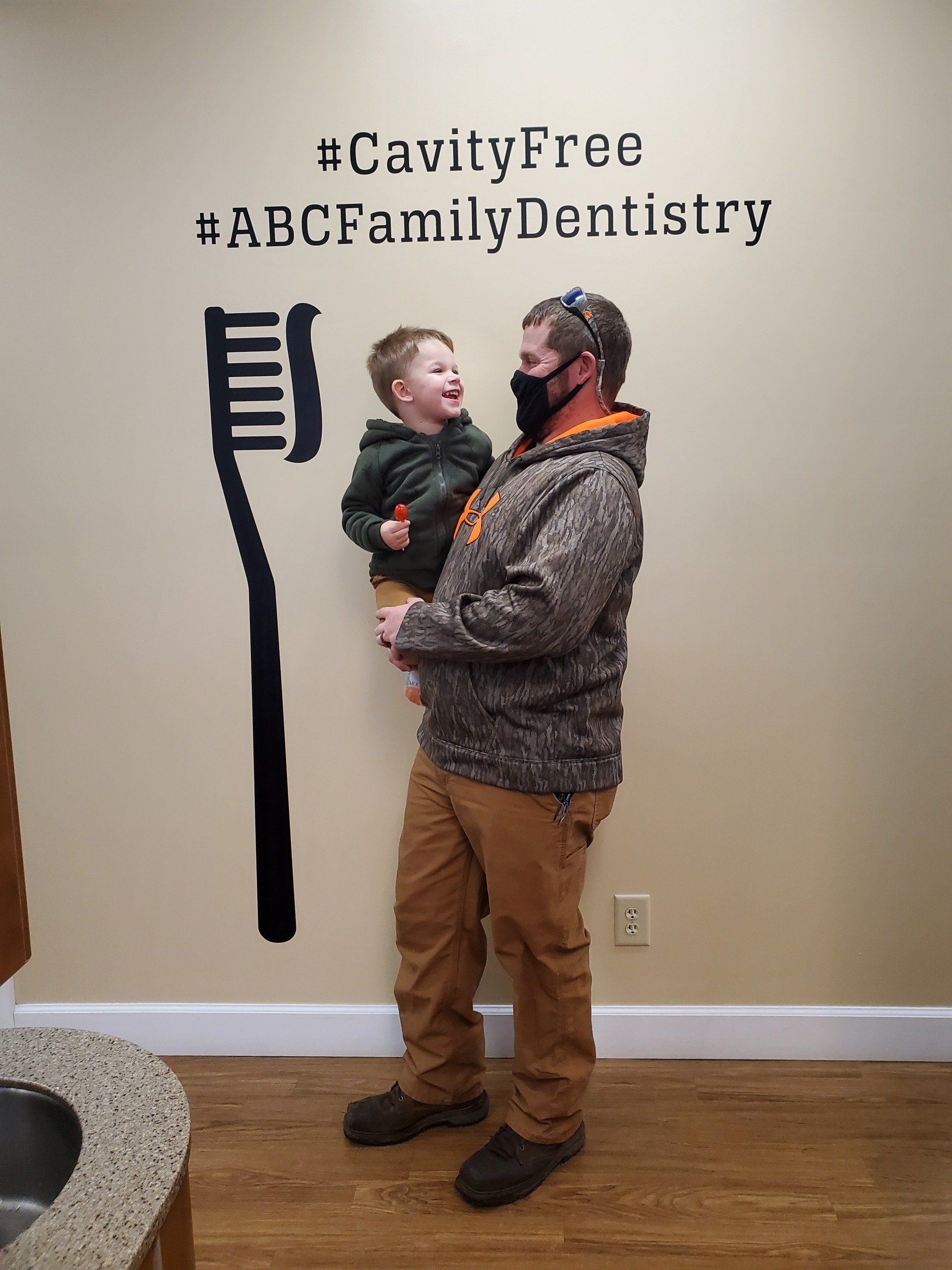 Man holding a happy child in front of a dental office wall with a large toothbrush and 
