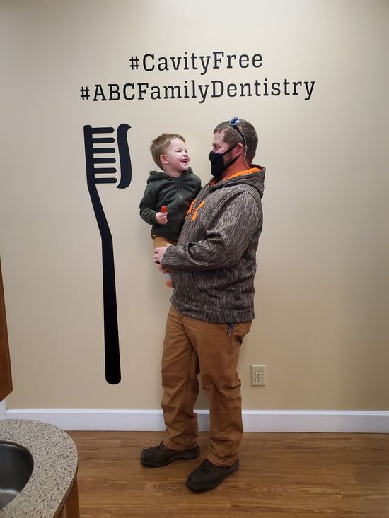 Man holding a happy child in front of a dental office wall with a large toothbrush and 