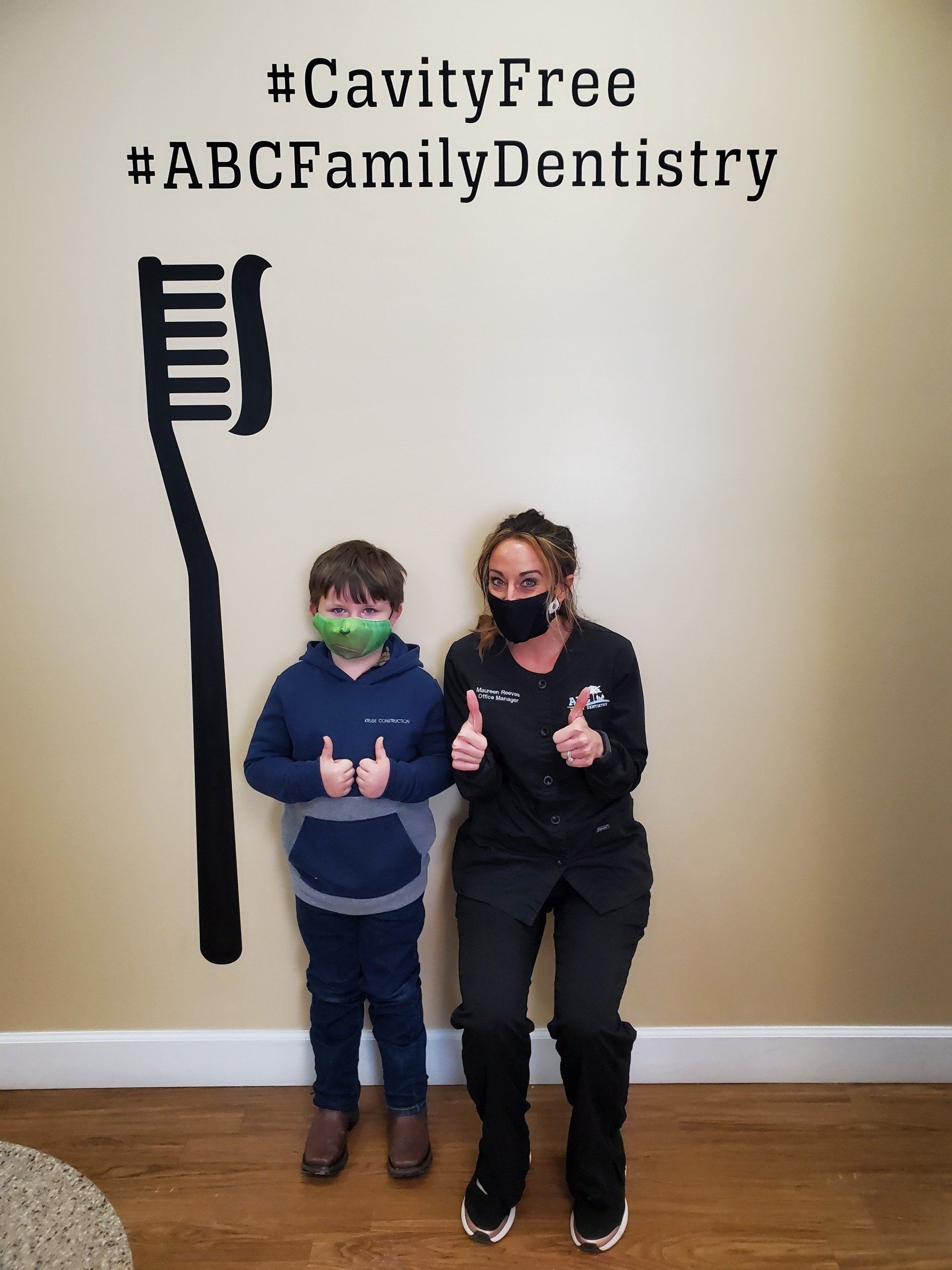 Boy and dentist giving thumbs up in front of a toothbrush wall graphic at ABC Family Dentistry. Both wear masks.