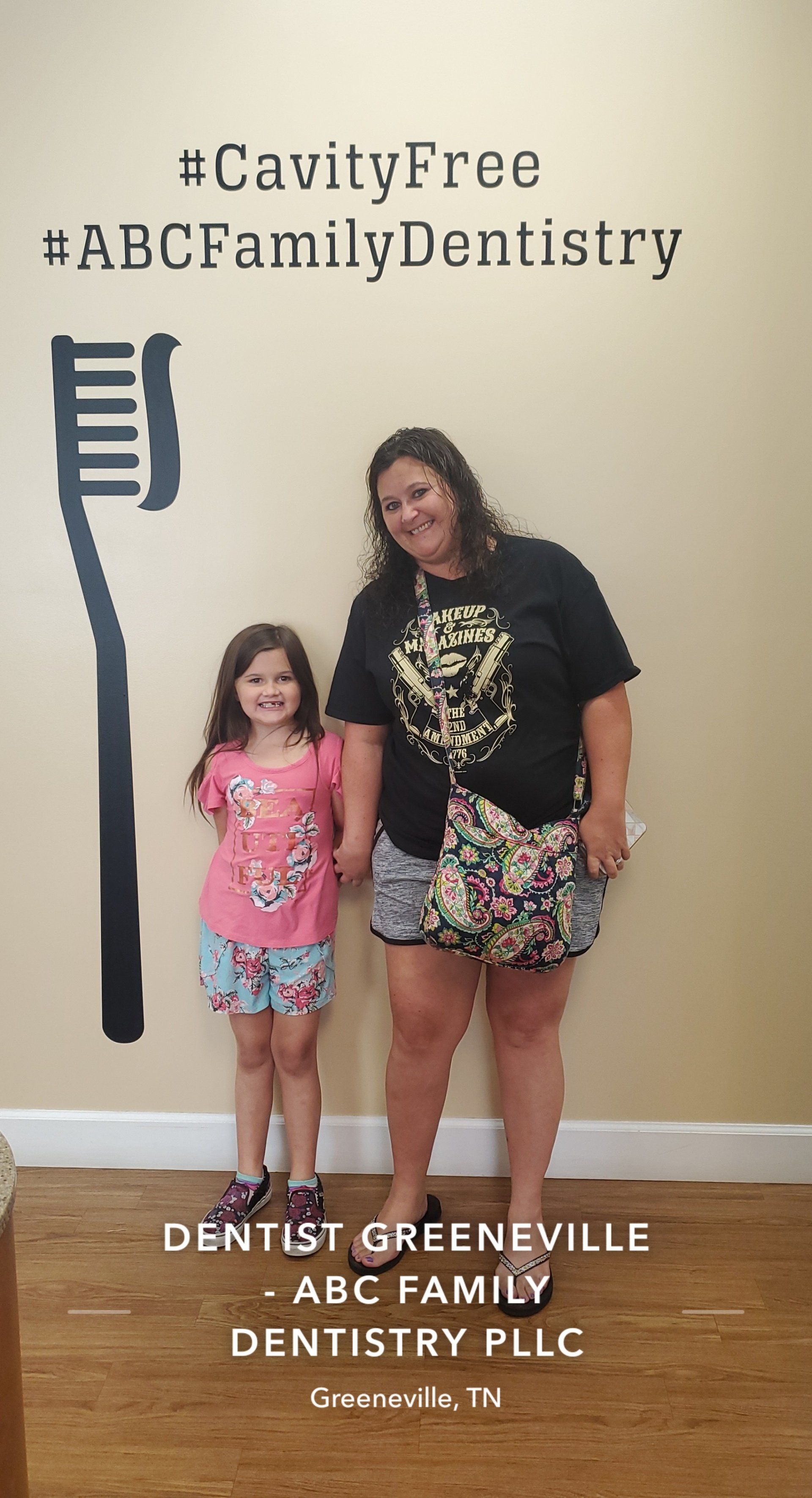 A woman and a girl smiling, holding hands, in front of a dentistry wall with a toothbrush graphic and hashtags.