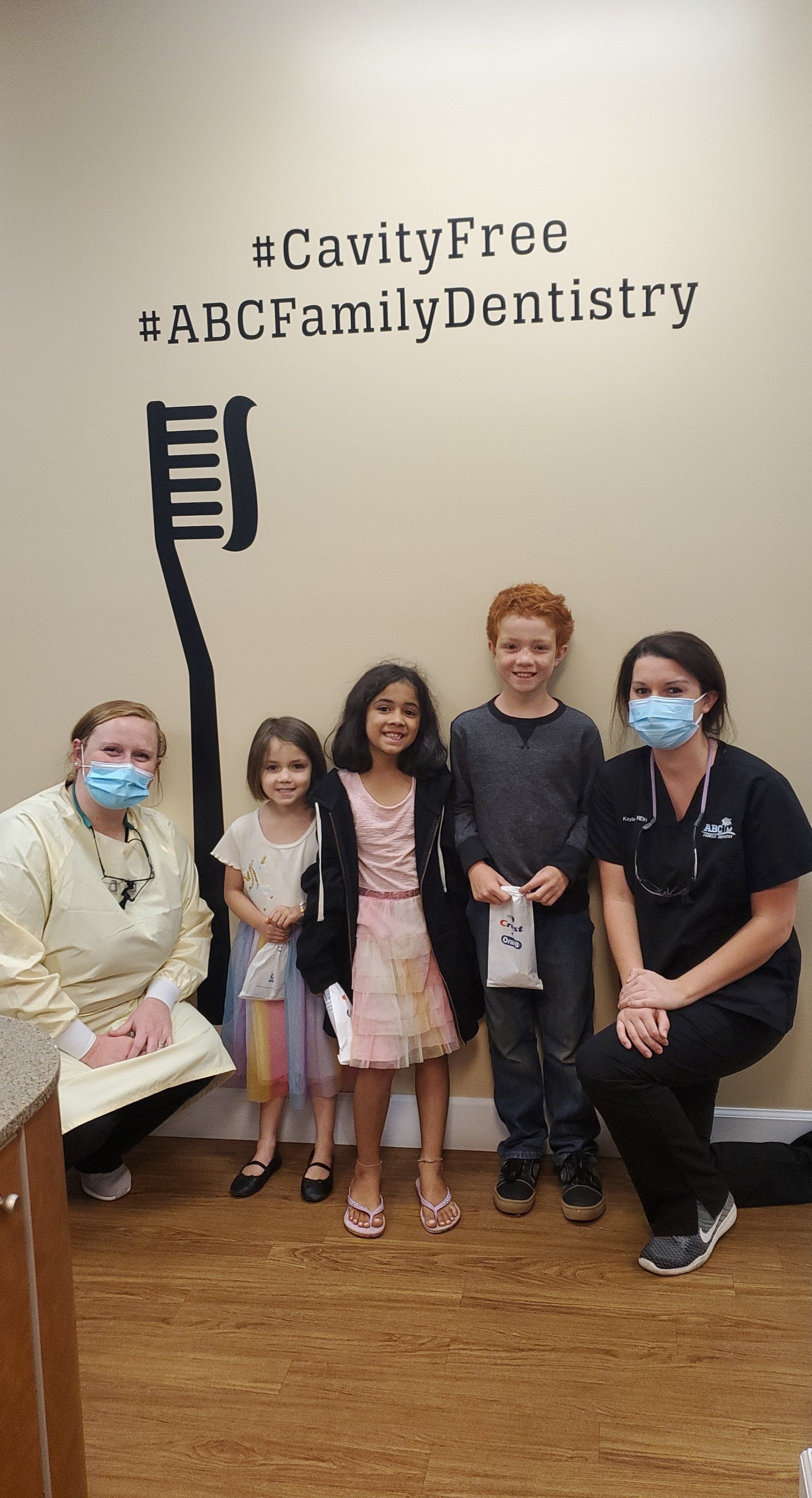 Two dental staff with masks pose with three smiling children in front of a wall with a toothbrush illustration and hashtags.