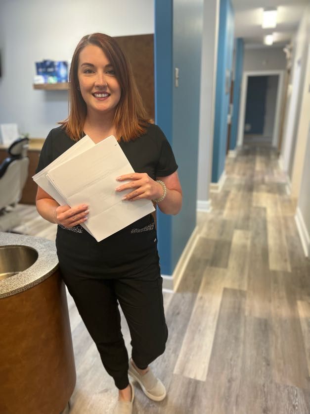 Woman in black scrubs holding papers in a dental office hallway, smiling at the camera.