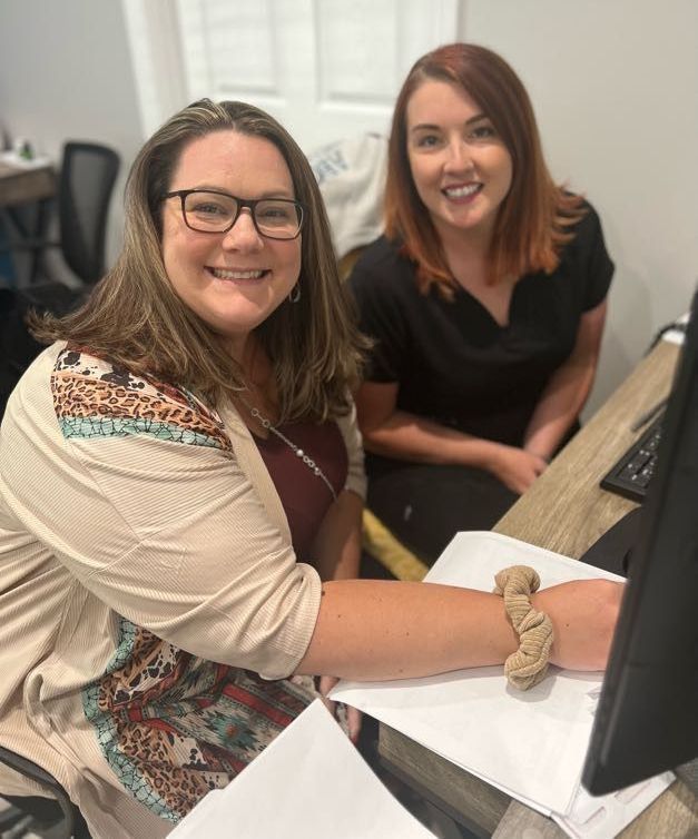 Two women smiling at a desk. One wears glasses and a tan cardigan, the other has red hair and a black shirt.